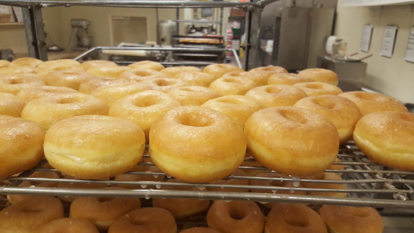 Trays of freshly fried donuts cooling on wire racks in the bakery at Ray's, with the mixer and work benches behind.