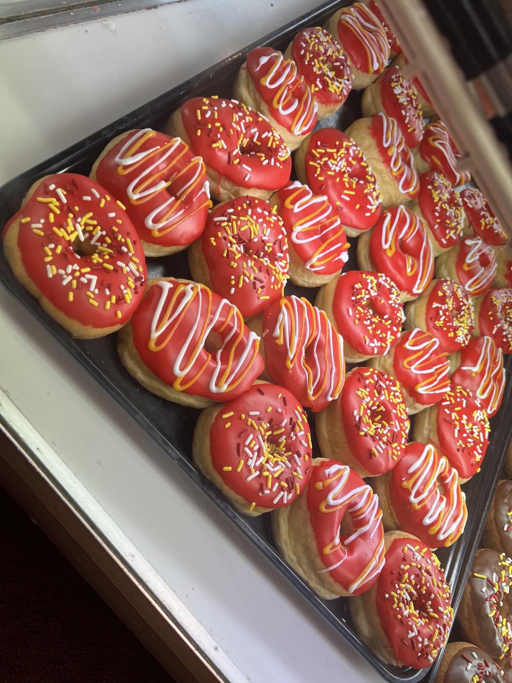 A full tray of donuts iced in red with red, yellow and white sprinkles and drizzle.